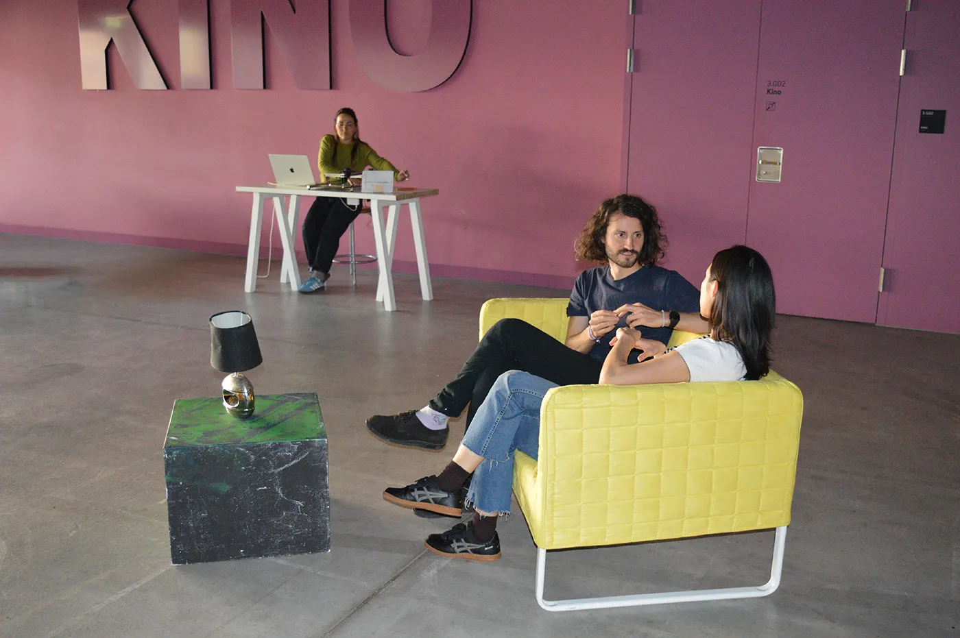 Two people sitting on a yellow sofa talking and person sitting in a table behind a pen-plotter