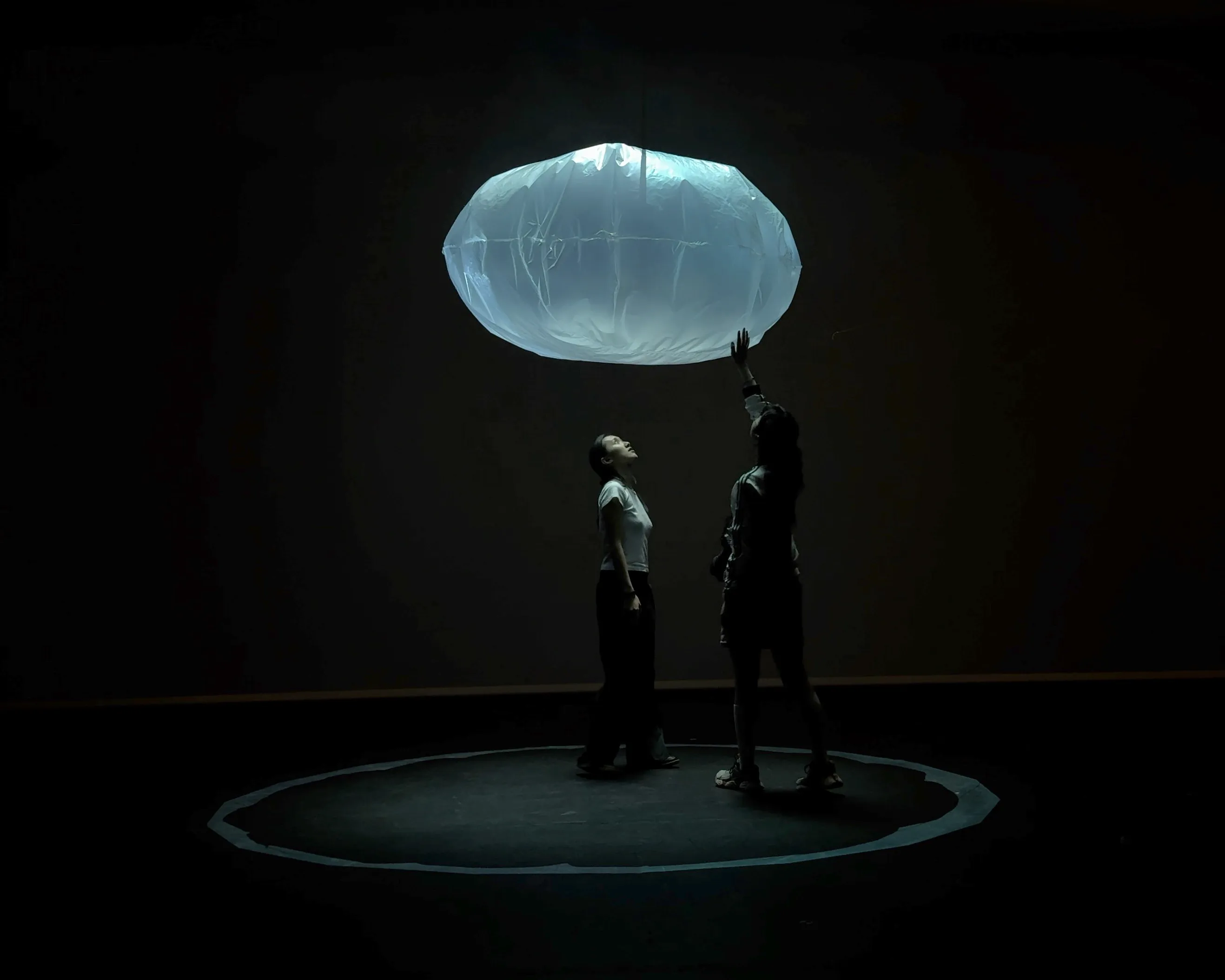 Two visitors standing beneath the floating bubble in a dark room, one reaching up to touch it