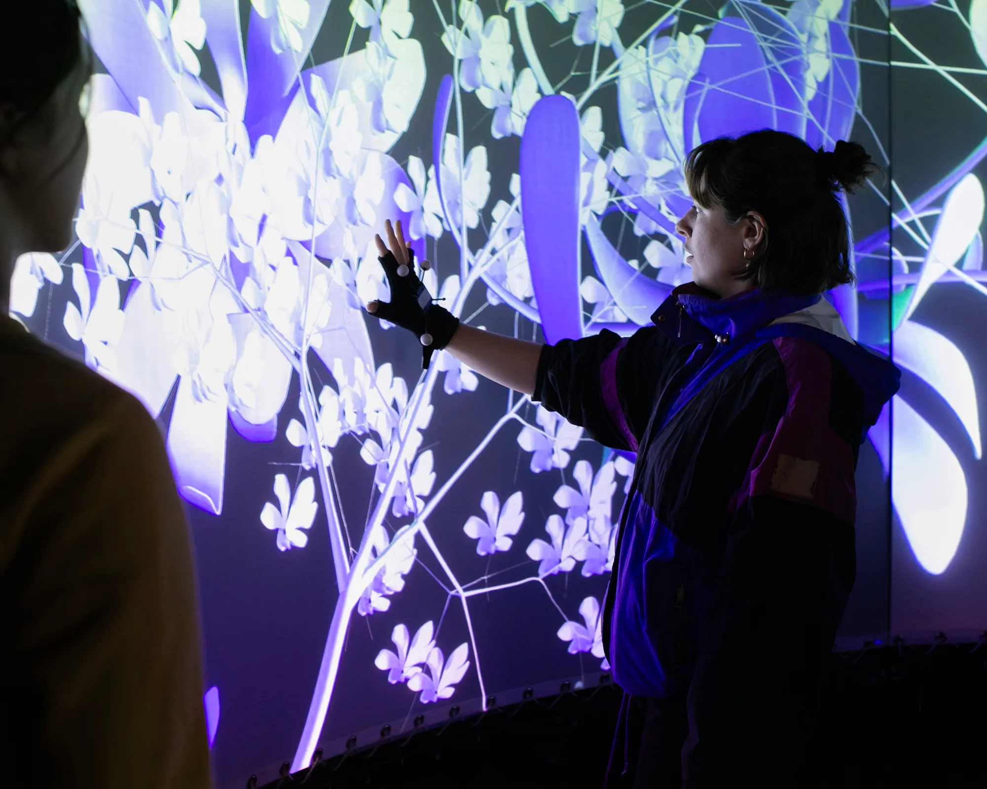 A visitor wearing a tracking glove reaching toward projected purple and white flowers